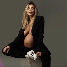 a pregnant woman sitting on top of a white stool wearing adidas shoes and posing for the camera