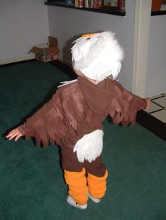 a small child dressed as an eagle standing on green carpeted floor in front of bookshelves