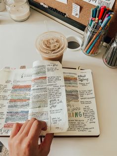 a person is writing on an open bible with colored pencils next to it and a cup of coffee in the background