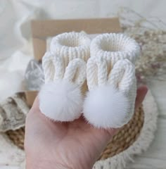 a hand holding two white knitted baby booties with pom - poms