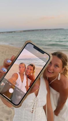 two women taking a selfie on their cell phone at the beach while wearing white dresses