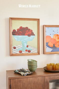 two paintings hanging on the wall next to a wooden cabinet with plates and bowls sitting on it