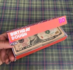 a person is holding a birthday dough box with money in it on a plaid tablecloth