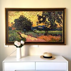 a painting is hanging on the wall above a white dresser with a hat and flowers