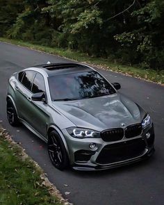 a silver bmw x6 driving down a road next to some green grass and trees