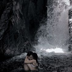 a woman sitting in the water next to a waterfall with her arms around her head