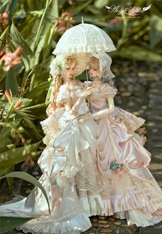 two dolls are sitting under an umbrella on the ground in front of some plants and flowers