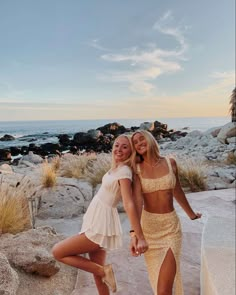 two women standing next to each other in front of rocks and the ocean at sunset