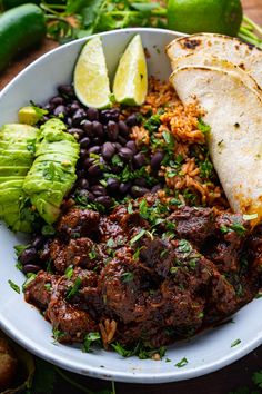 a white plate topped with meat, beans and tortillas next to lime wedges