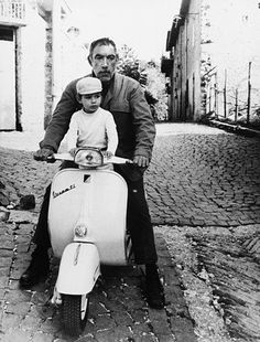 a man and child riding on the back of a scooter down a cobblestone street