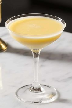 a glass filled with yellow liquid sitting on top of a white marble table next to a bottle