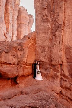 a bride and groom standing in front of large rocks at the base of a cliff