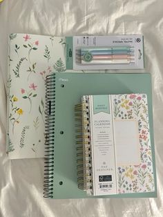 two notebooks and a pen sitting on top of a white sheet with floral designs