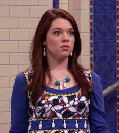 a woman with red hair wearing a blue top and earrings standing in front of a tiled wall