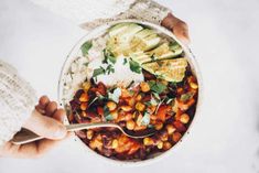 two hands holding a bowl of food with beans, avocado and sour cream