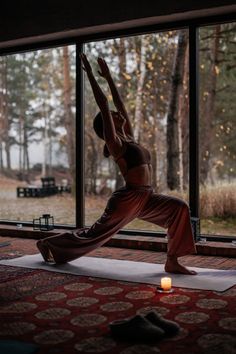 a woman practices yoga in front of a large window with a candle on the floor