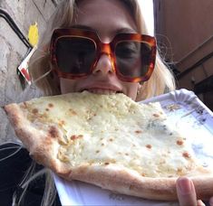 a woman wearing sunglasses eating a large piece of pizza in front of her face and looking at the camera