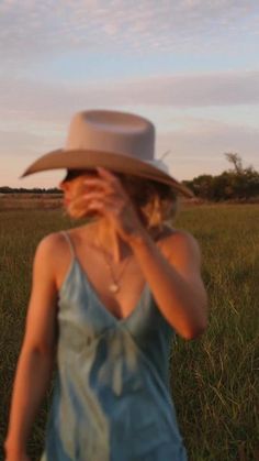 a woman in a blue dress and cowboy hat walking through the grass with her hands to her face