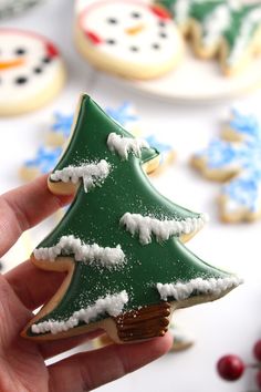 a hand holding a decorated christmas tree shaped cookie in front of snow - covered cookies