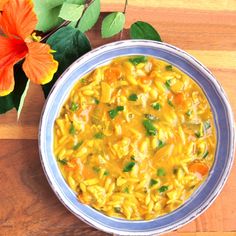 a bowl of pasta soup next to an orange flower on a wooden table with green leaves