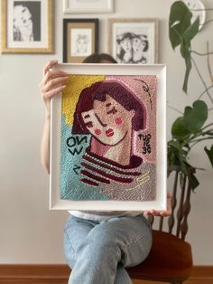 a person sitting on a chair holding up a framed art piece with an image of a woman's face