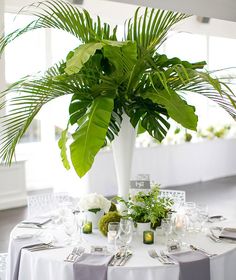 there is a large plant in the center of this table with white linens and place settings