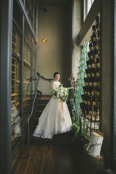 a woman in a wedding dress is standing on the stairs with her bouquet and wine bottles behind her