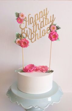 a white cake topped with pink flowers and a gold happy birthday sign in the middle