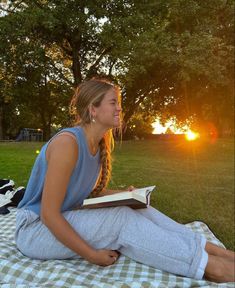 a young woman sitting on top of a blanket reading a book in the park at sunset