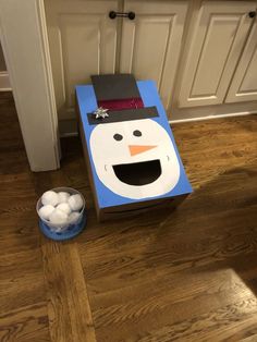 a cardboard snowman box sitting on top of a wooden floor next to a bowl of white balls