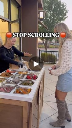 two women standing in front of a counter filled with food and talking to each other