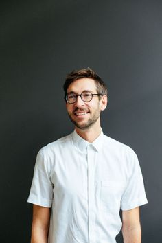 a man wearing glasses standing in front of a black wall with his hands in his pockets