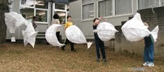 several people holding up white kites in front of a building with trees and leaves on the ground