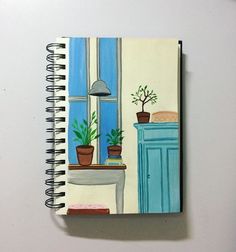 an open notebook with a painting of a desk and potted plants on the table