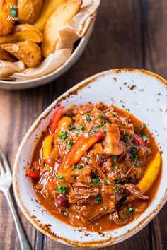 a white bowl filled with meat and vegetables next to a plate of fried potatoes on a wooden table