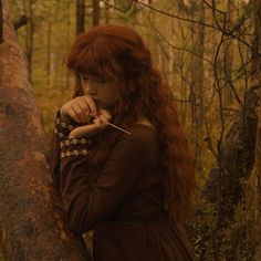 a woman with long red hair is leaning on a tree branch in the woods looking at something