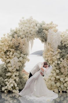 a bride and groom kissing in front of an arch of flowers with the ocean in the background