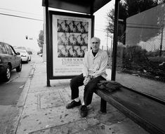 an older man sitting on a bench in front of a bus stop with a movie poster behind him