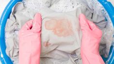 two hands in pink gloves washing clothes in a blue tub with foam on the floor