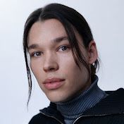 a man with long hair wearing a black jacket and looking at the camera while standing in front of a white background