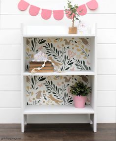 a white book shelf with pink bunting on the top and wallpaper behind it