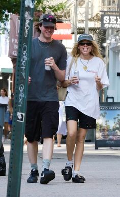 a man and woman walking down the street holding drinks in their hands while wearing hats