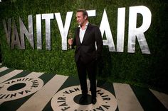 a man in a tuxedo standing next to a vanity fair sign on the red carpet