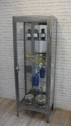 a glass case with some bottles and other items in it on a wooden floor next to a white brick wall