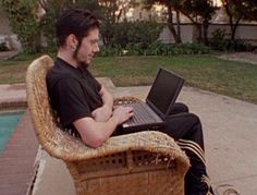 a man sitting on a wicker chair with his laptop in front of the pool