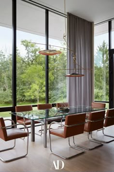 a glass dining table surrounded by brown chairs in front of large windows with trees outside