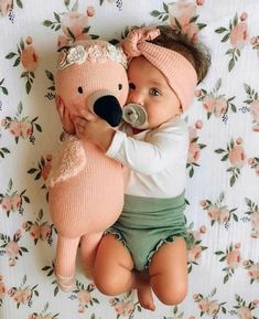 a baby laying on top of a bed next to a stuffed teddy bear wearing a pink hat