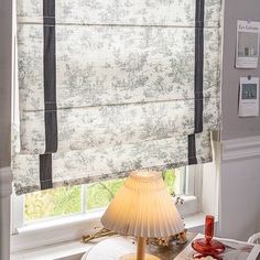 a lamp sitting on top of a wooden table next to a window covered in roman shades