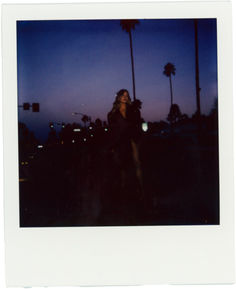 a polaroid photograph of a woman standing in front of palm trees at night time