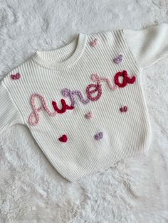 a white sweater with the word mom written in pink and red hearts on it, sitting on a fluffy surface